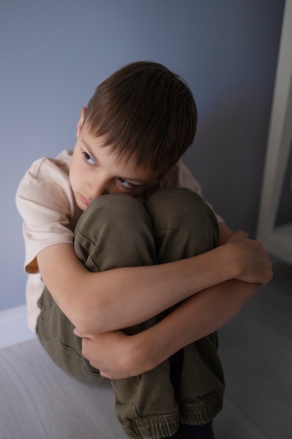 The image shows a young boy sitting on the floor in a corner, hugging his knees to his chest. He has a sad or contemplative expression, with his gaze directed downward and to the side. His body language suggests feelings of loneliness, anxiety, or deep thought. The setting appears to be indoors, with a grayish-blue wall in the background and soft, natural lighting. The overall mood of the image conveys a sense of isolation or introspection.