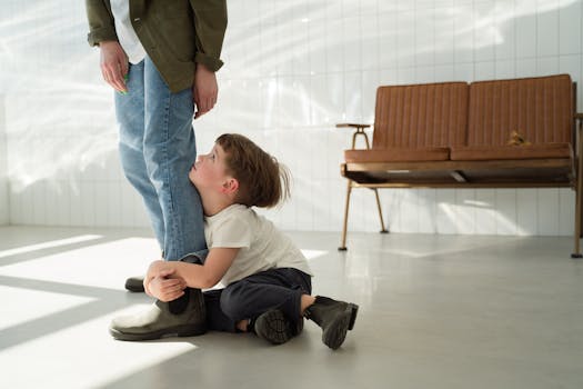 The image shows a young child sitting on the floor, clinging tightly to an adult’s leg. The child, wearing a white shirt and dark pants, looks up at the adult with an expression that suggests a need for comfort, security, or attention. The adult, dressed in casual attire with blue jeans and a green jacket, stands still, with only their lower body visible. The background features a bright, minimalist space with white tiled walls and a vintage brown couch, creating a contrast between the warm sunlight and the emotional tone of the scene. The image conveys themes of attachment, dependence, or a moment of emotional connection.